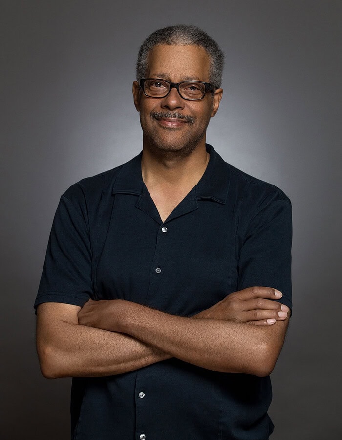 Casual studio portrait of a man wearing glasses and a dark short-sleeve shirt with folded arms.