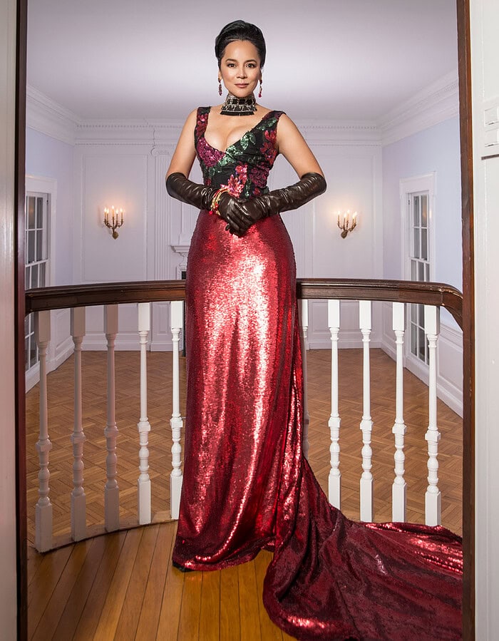 Publicity portrait of a woman in a red sequined gown standing in an elegant white interior.