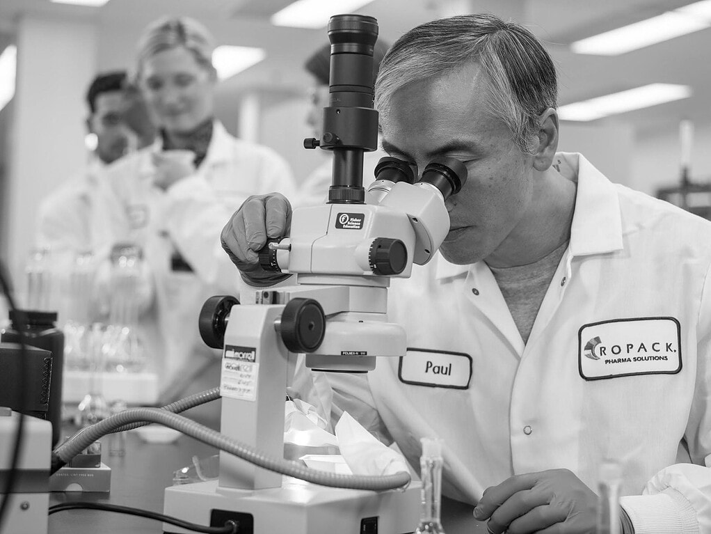Scientist looking through a microscope in a pharmaceutical laboratory while colleagues work behind him.