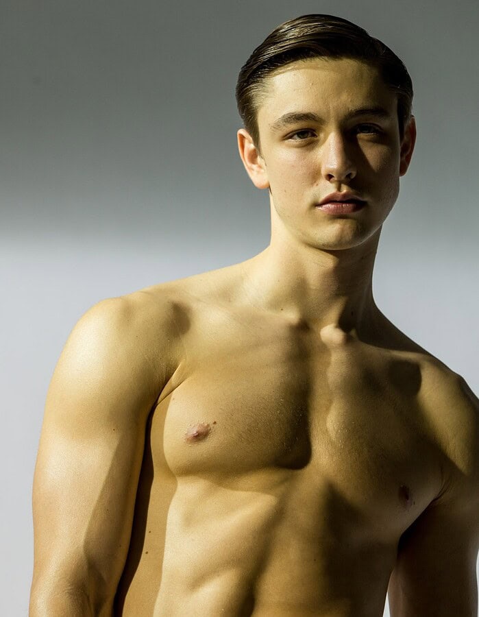 Studio portrait of a shirtless young male model lit with dramatic directional light against a gray background.