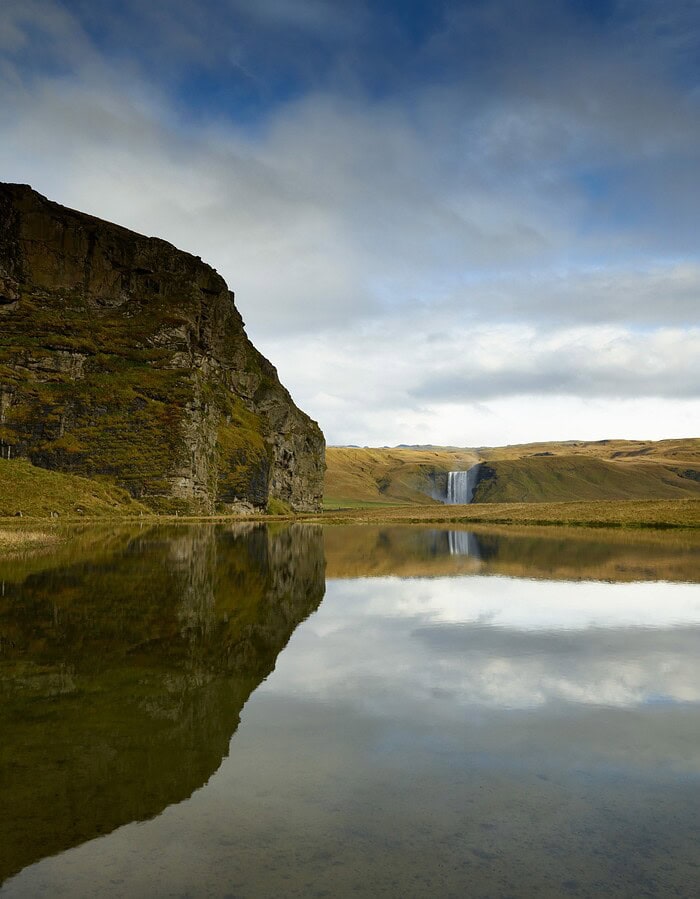 Distant Iceland waterfall reflected in calm water beside a steep green cliff.
