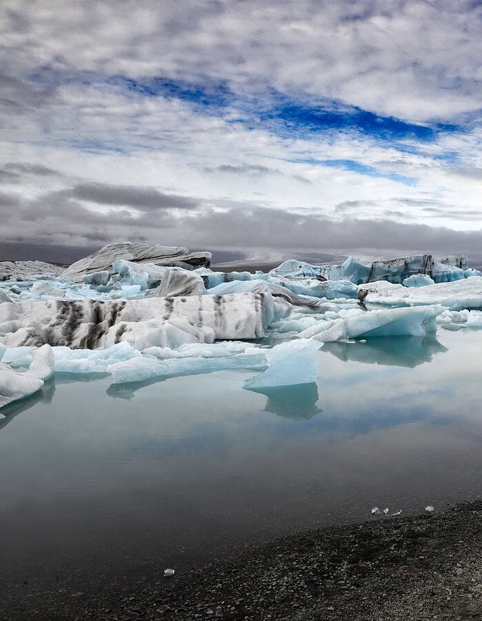 Wide view of an Iceland glacier lagoon with blue icebergs, reflections, and a small figure on shore.