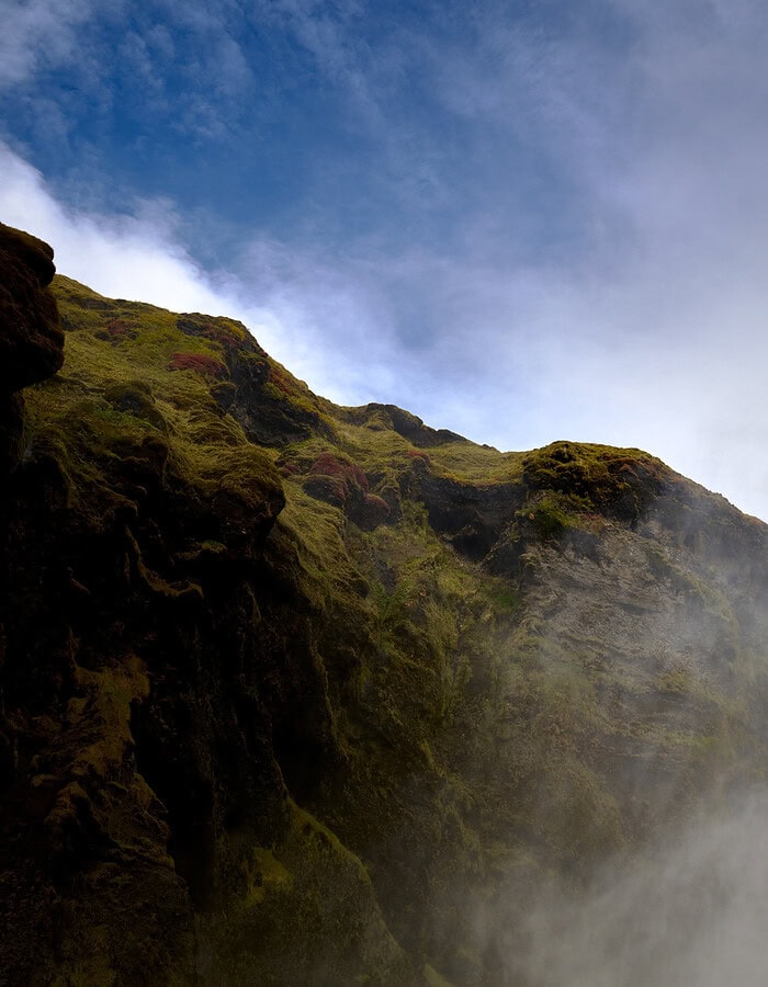 Powerful Iceland waterfall falling between dark cliffs and mist under a blue sky.