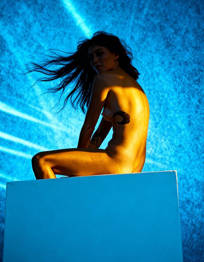 Implied nude figure study with blue background, warm light, and windblown hair.