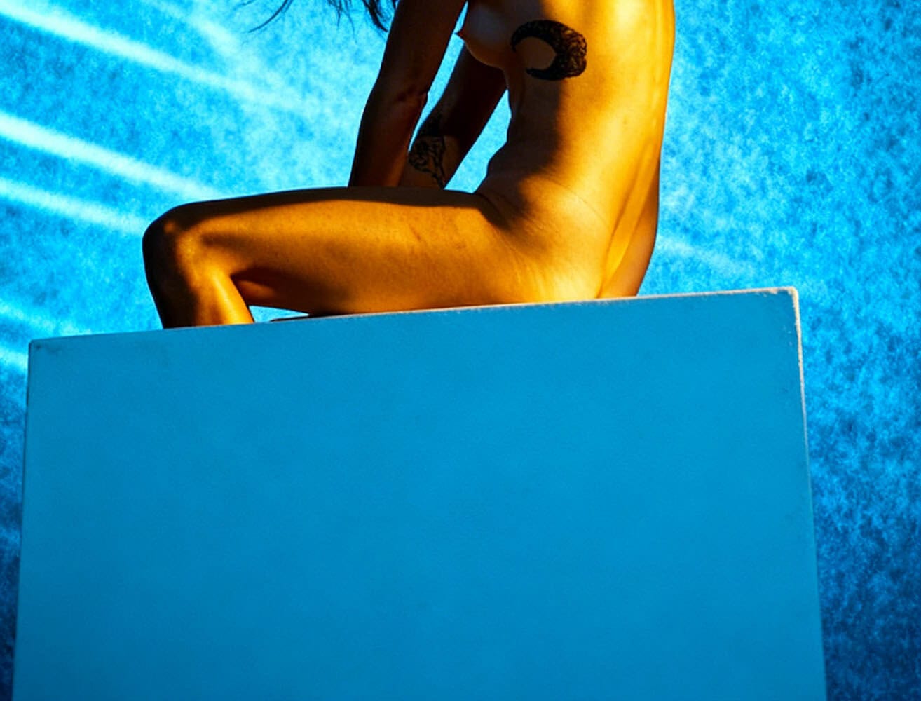 Implied nude figure study with blue background, warm light, and windblown hair.