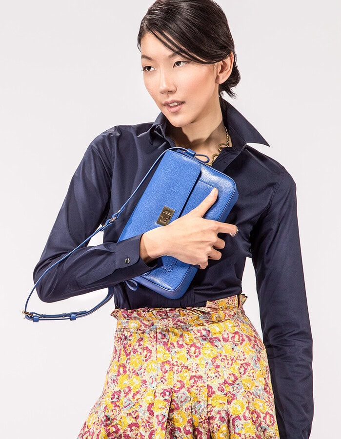 Standing fashion portrait of a model holding a blue leather handbag with a navy blouse and patterned skirt.