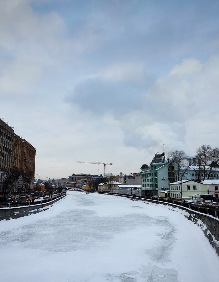 Snow-covered frozen river or canal running through Moscow beneath a pale winter sky.