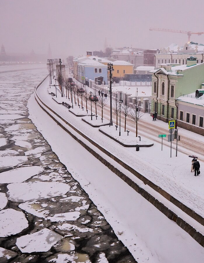 Snowy Moscow riverside street with pastel buildings and ice floating on the water.
