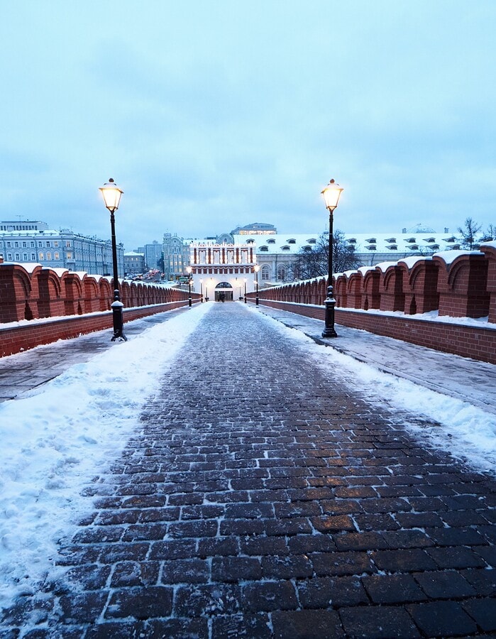 Snow-covered Moscow bridge leading between red brick walls and glowing lamps at dusk.