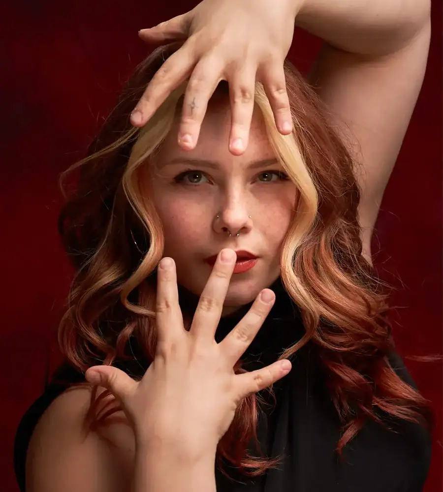 Close portrait of a dancer with wavy hair and facial piercings, framed by hands against a red backdrop.