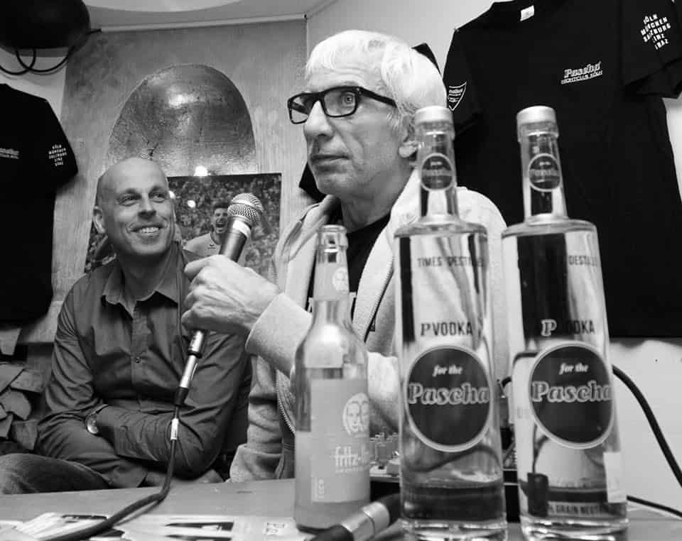 Schwarzweißfoto zweier Männer, einer mit Mikrofon, auf dem Tisch Flaschen mit der Aufschrift „Pascha"