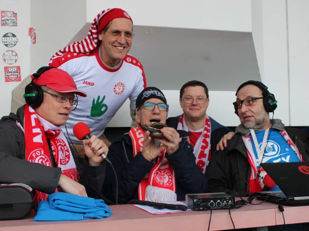 Fünf Männer mit Kopfhörern und Mikrofonen bei einer Radioübertragung, alle mit rot-weißen FC-Köln-Schals