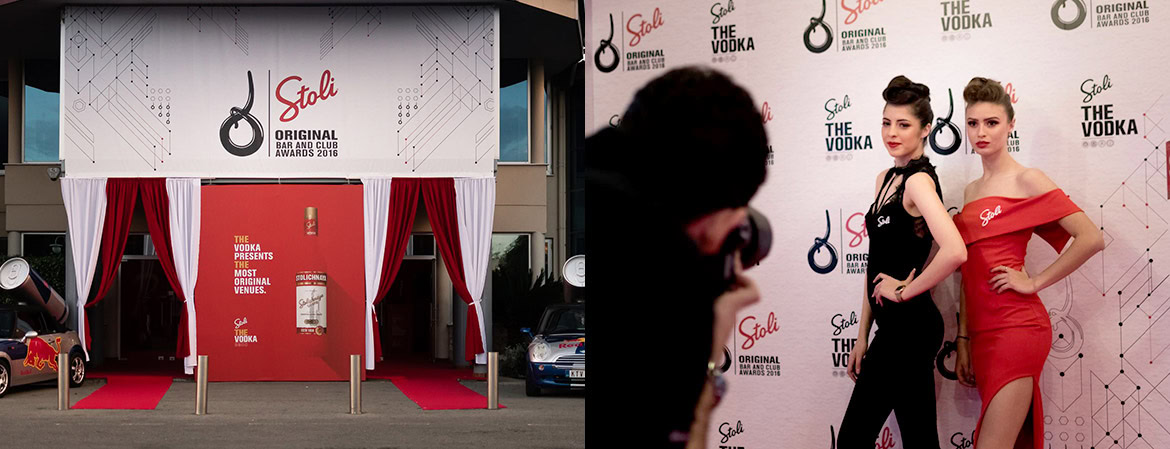 A red carpet entrance with white curtains and a backdrop featuring logos, with two women posing for photos in front of it.