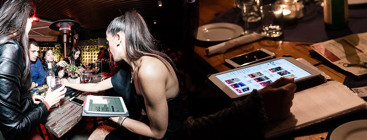 A group of people sitting at a bar, using tablets and smartphones, with drinks and menus on the table.