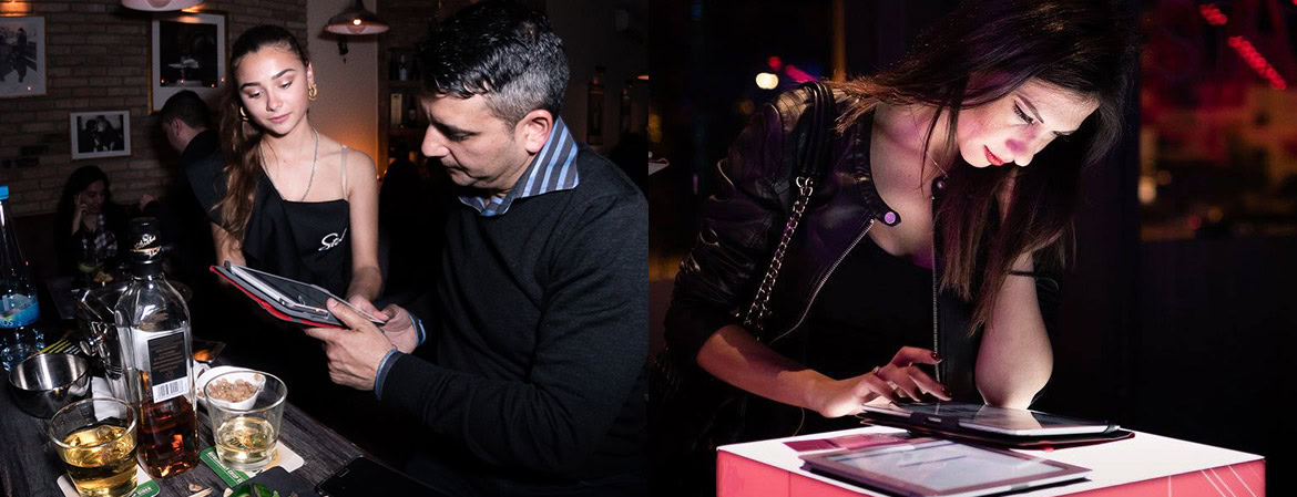 A group of people socialising at a bar, with drinks and a woman using a tablet.