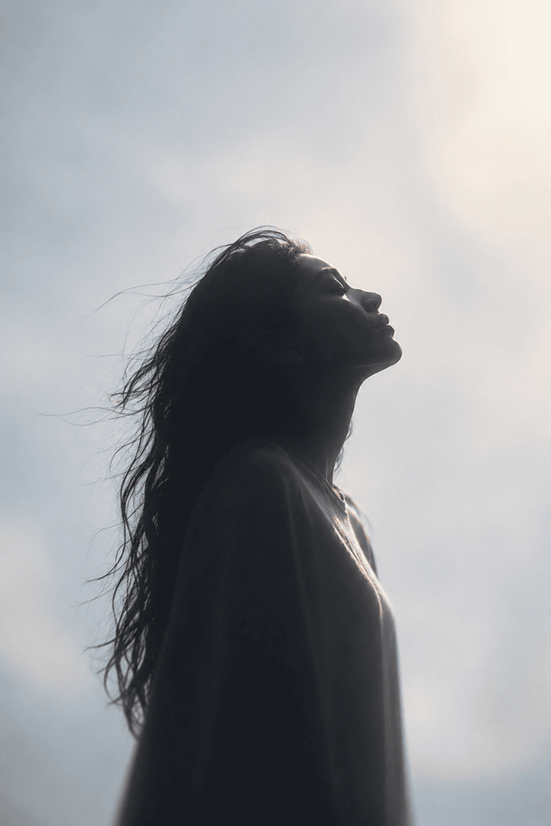 A woman with long hair facing the sky, backlit by sunlight, with clouds in the background.