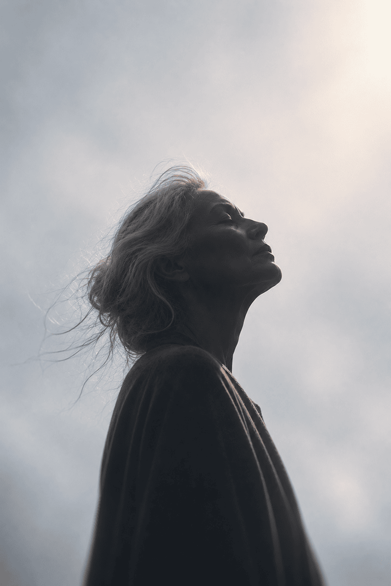 A woman with grey hair looking up against a cloudy sky with sunlight.