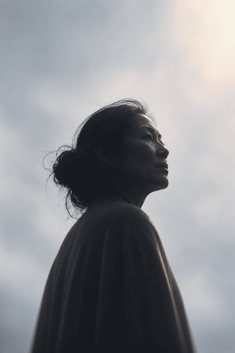 A woman with dark hair in a bun looks to the side against a cloudy sky background.