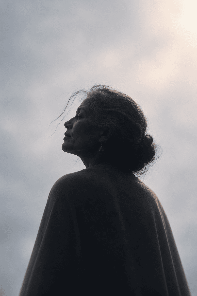 A woman with dark hair and a dark coat looks to the sky against a cloudy background.