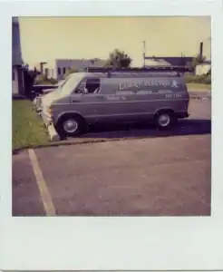 A gray van labeled "Lowry Electric" is parked in a lot beside a grassy area, with buildings visible in the background, offering a glimpse into what you might see when learning more about us.