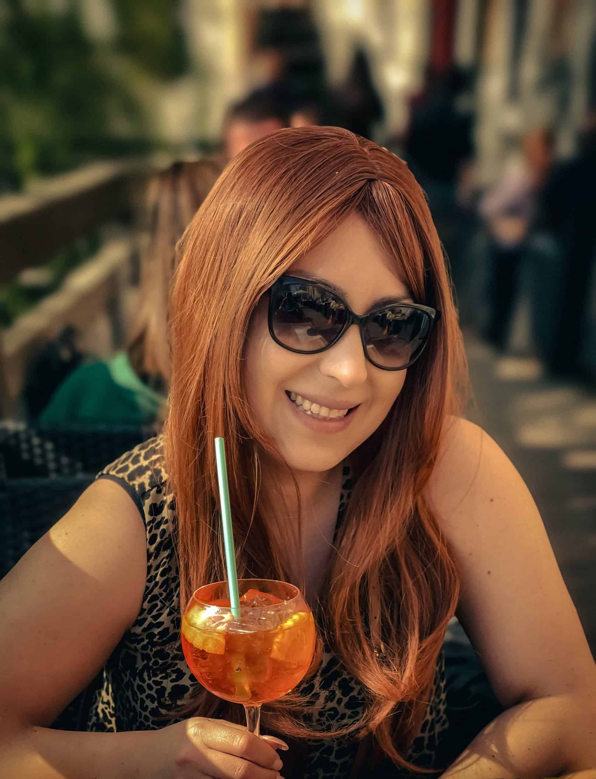 Woman with red hair and sunglasses enjoying a cocktail outdoors.