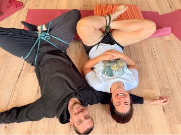 Zwei Personen liegen auf einem Holzboden, die Beine mit Seilen zusammengebunden, und lächeln in die Kamera. Sie sind leger gekleidet und von Yogamatten und einem Kissen umgeben - vielleicht machen sie eine spielerische Pause während ihrer NLP-Ausbildung.