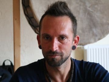 Ein Mann mit kurzen dunklen Haaren und Messgeräten in den Ohren schaut in die Kamera, er trägt ein dunkles Hemd und eine Halskette. Hinter ihm hängt ein runder Gegenstand an der Wand - vielleicht ist er bereit, seine NLP-Ausbildung oder seine Reise als Coach zu beginnen.