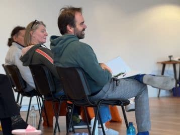 Eine Gruppe von Personen sitzt in einem Raum auf Stühlen und blickt aufmerksam nach vorne. Eine Person, die vielleicht gecoacht werden will, hält Papiere in der Hand und hat ein Bein über das andere geschlagen - vielleicht während einer NLP-Ausbildungssitzung.