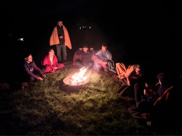 Eine Gruppe von Menschen sitzt nachts um ein Lagerfeuer, einige in Decken gehüllt, eine Person spielt auf der Gitarre, erzählt von ihrer NLP-Ausbildung und genießt die Wärme ihrer Mitgliedschaft.