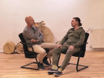 Zwei Männer sitzen sich auf Stühlen gegenüber und unterhalten sich in einem Raum mit Holzboden und Trommeln im Hintergrund darüber, wie man Coach wird.