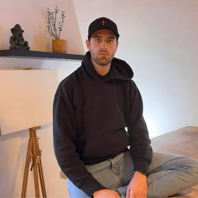 Ein Mann in schwarzem Kapuzenpulli und Mütze sitzt an einem Holztisch in einem Raum mit hellen Wänden, einem Regal mit Dekoration und einer leeren Leinwand auf einer Staffelei.