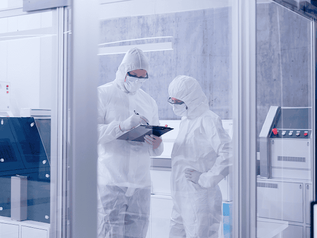 Two technicians in cleanroom suits reviewing notes on a clipboard inside a controlled laboratory environment.
