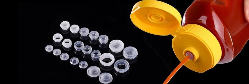 Assorted pink, white, and clear plastic bottle caps alongside a red safety squeeze bottle with yellow cap and orange nozzle, placed on black background.