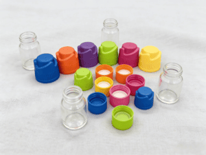 Assorted colorful powder dispensing caps and small glass jars on a white surface, showcasing innovative packaging solutions for modern products.