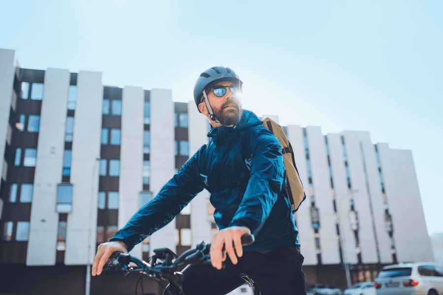 Mann mit Fahrrad, Fahrradhelm, Sonnenbrille vor moderner Gebäudefront.