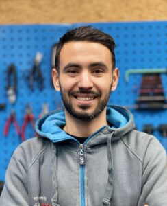 Rad & Tat Tübingen Werkstatt Mann in Werkstatt vor blauer Werkzeugwand, lächelnd.