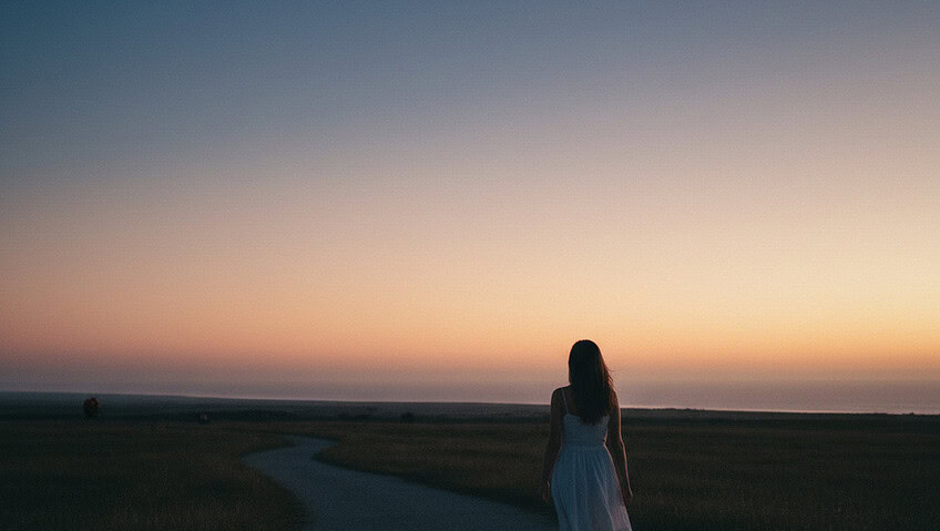 Woman in a white dress walks along a winding path at dusk toward a colorful sunset over open fields.