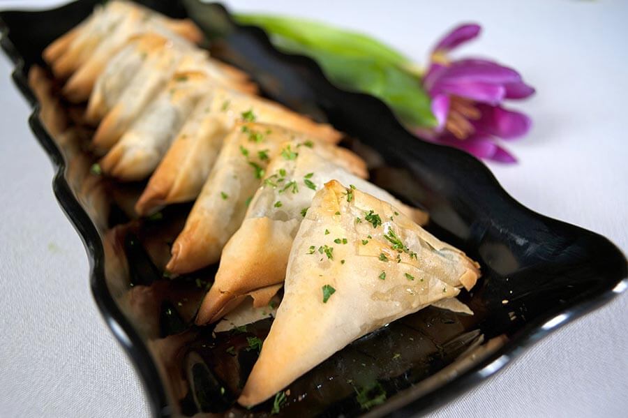 Golden spanakopita triangles garnished with fresh herbs on a wavy black platter next to a purple flower.