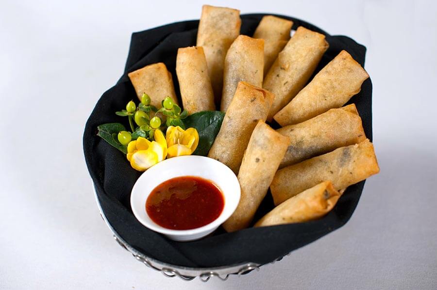 Crispy golden spring rolls served in a basket lined with a black napkin, accompanied by a small bowl of sweet and sour dipping sauce and a yellow flower garnish.