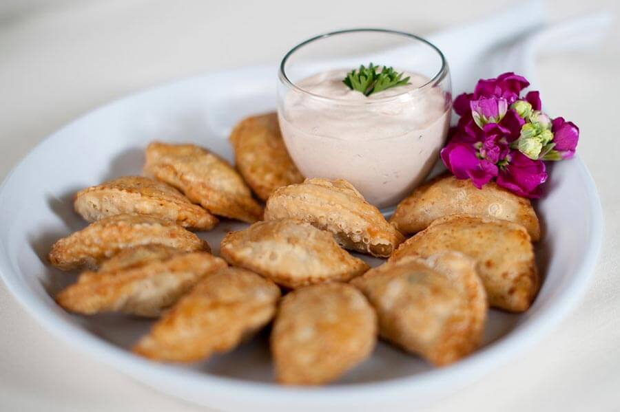 Mini empanadas or turnovers arranged on a white platter with a creamy dipping sauce and purple flowers.
