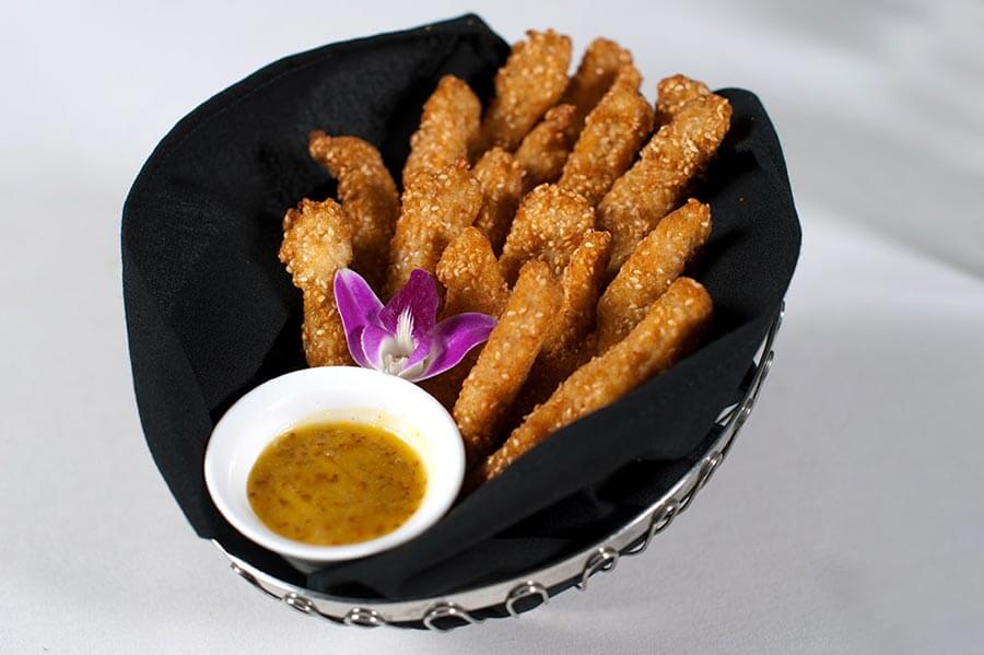 Sesame-crusted chicken tenders served in a black-lined basket with a side of honey mustard dipping sauce and a purple orchid garnish.