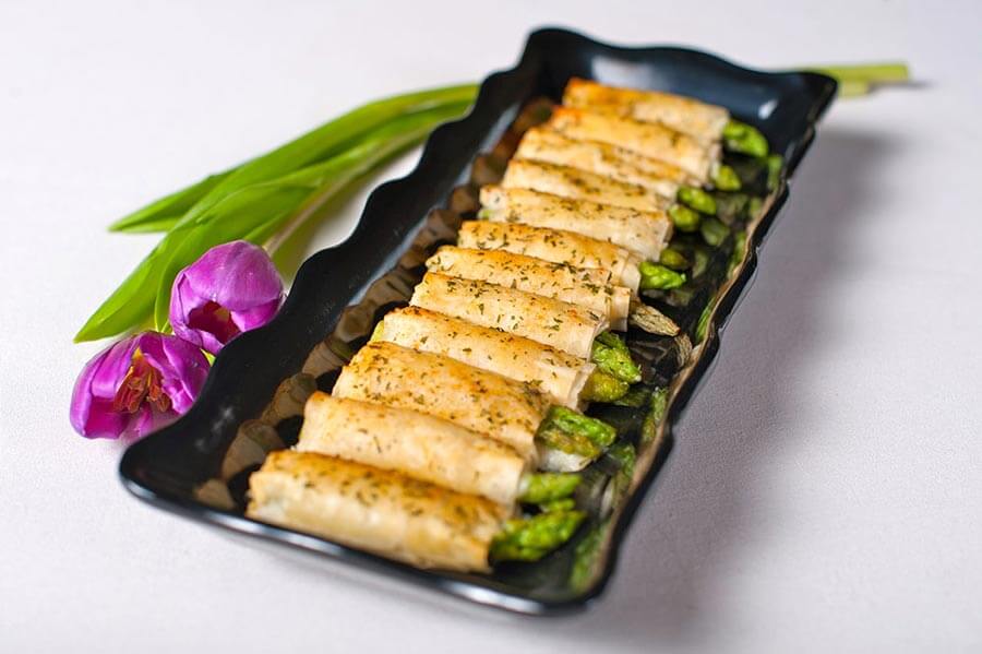 Baked asparagus pastry wraps sprinkled with herbs lined up on a black serving platter next to purple tulips.