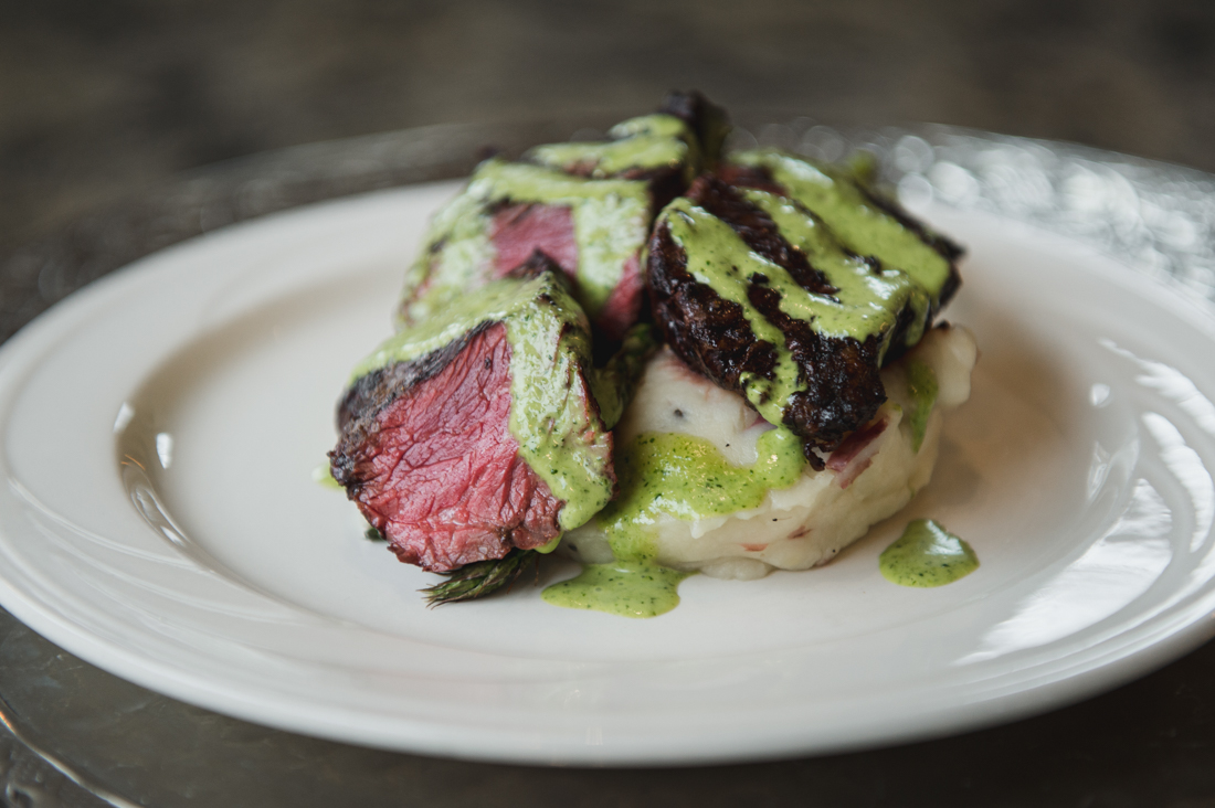 Sliced medium-rare steak over mashed potatoes, drizzled with a bright green herb sauce.