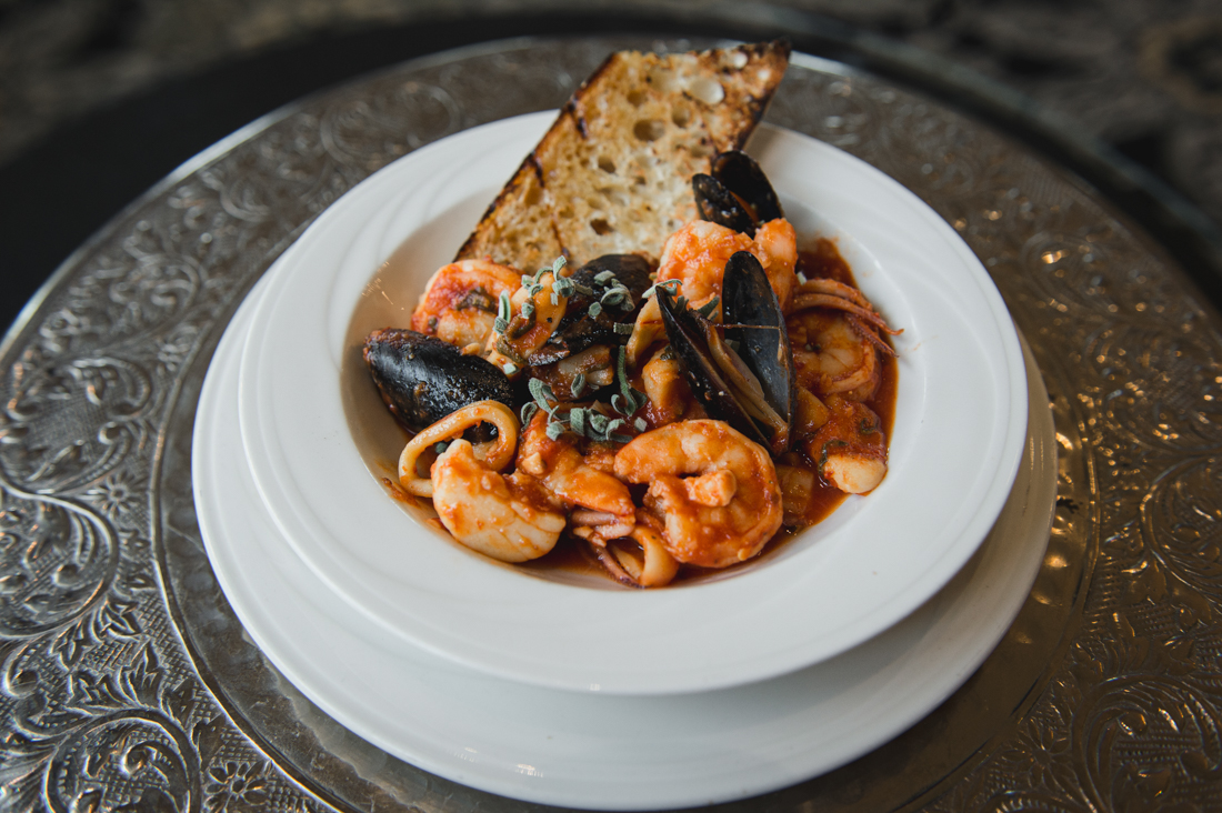 Seafood cioppino or stew filled with shrimp, mussels, and calamari in a tomato broth, served with a piece of grilled bread in a white bowl.
