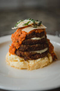 Baked eggplant parmesan tower layered with melted cheese, served over polenta or mashed potatoes, topped with marinara sauce, shaved cheese, and fresh basil.