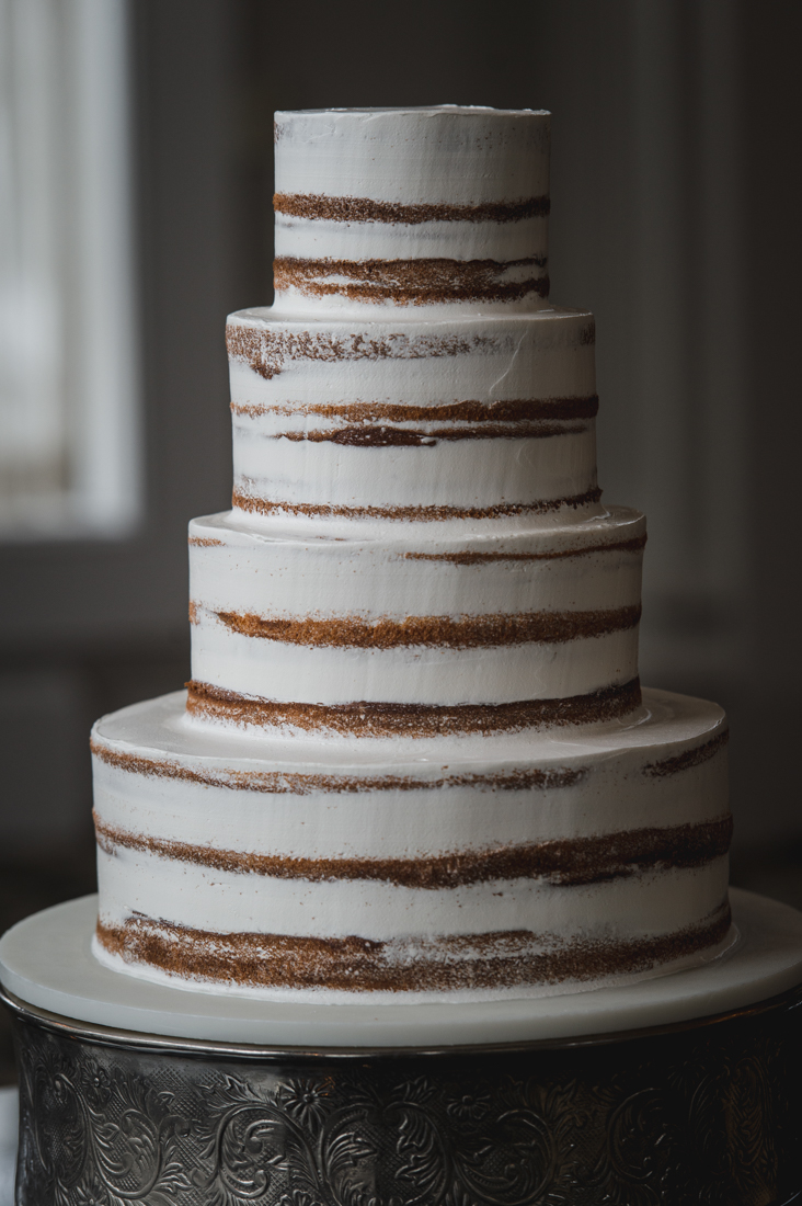 Four-tier semi-naked wedding cake with horizontal stripes of exposed cake layers showing through thin white frosting, displayed on a silver cake stand.