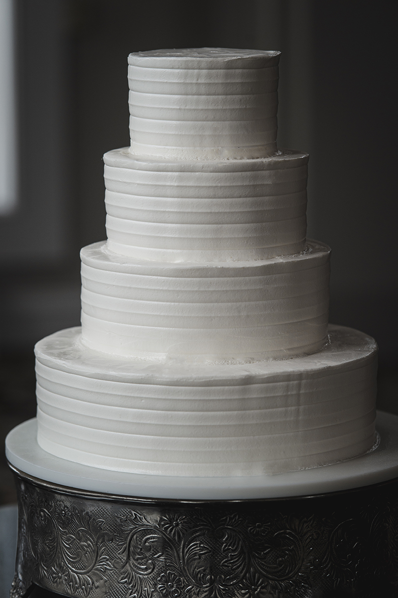 Four-tier plain white wedding cake with horizontal textured ridges on an ornate silver stand.