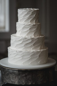 Four-tier white wedding cake with textured petal buttercream frosting on an ornate silver stand.
