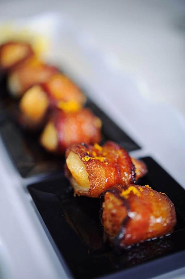 Bacon-wrapped sea scallops garnished with citrus zest and served on individual small black plates.