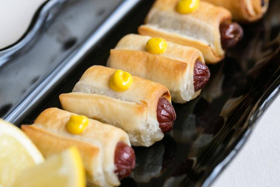 Pigs in a blanket with mustard drops lined up on a black serving platter.